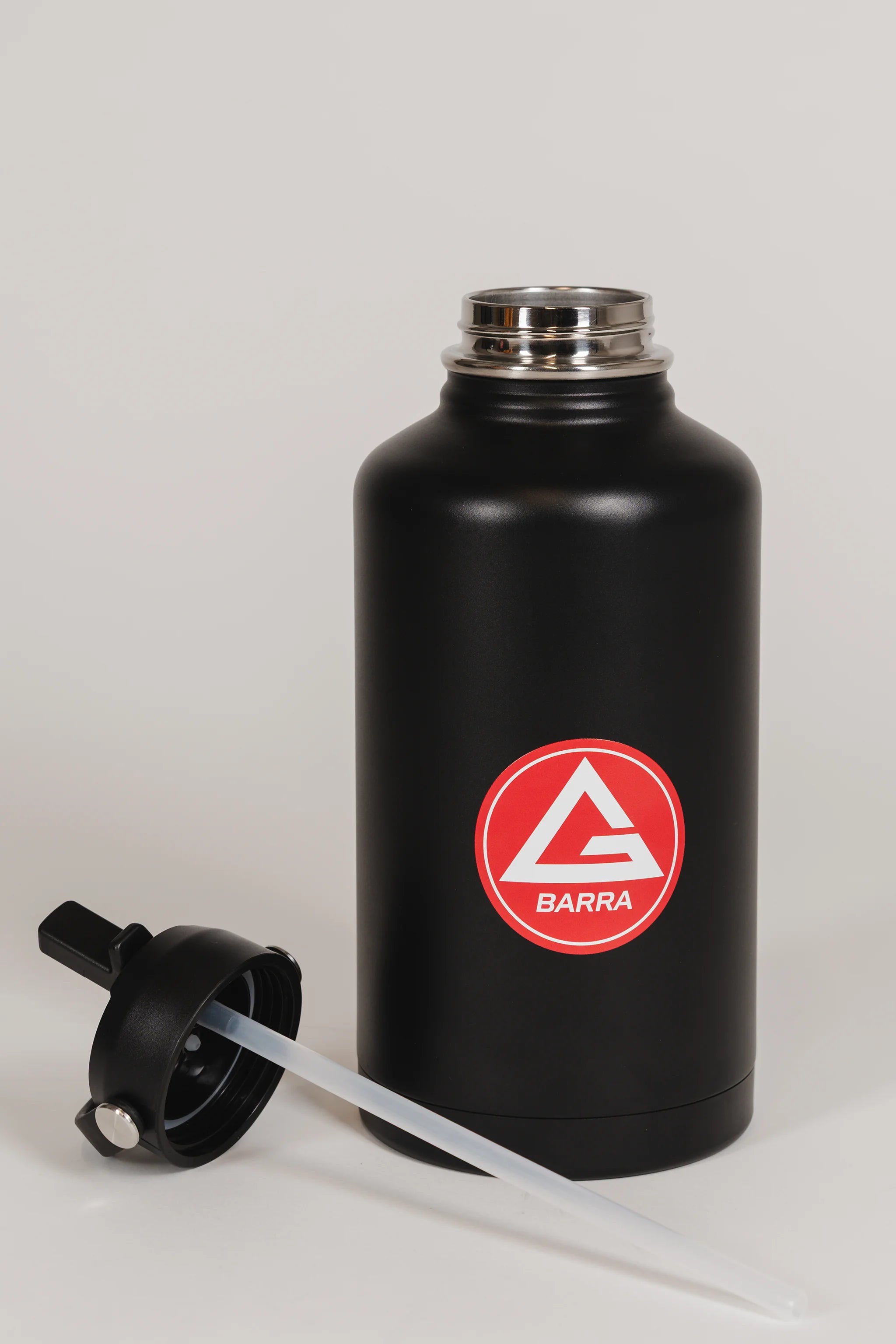 Black stainless steel 64 oz Red Shield bottle with the Gracie Barra Red Shield logo, shown with the straw lid removed and placed in front of the bottle, photographed against a light background.