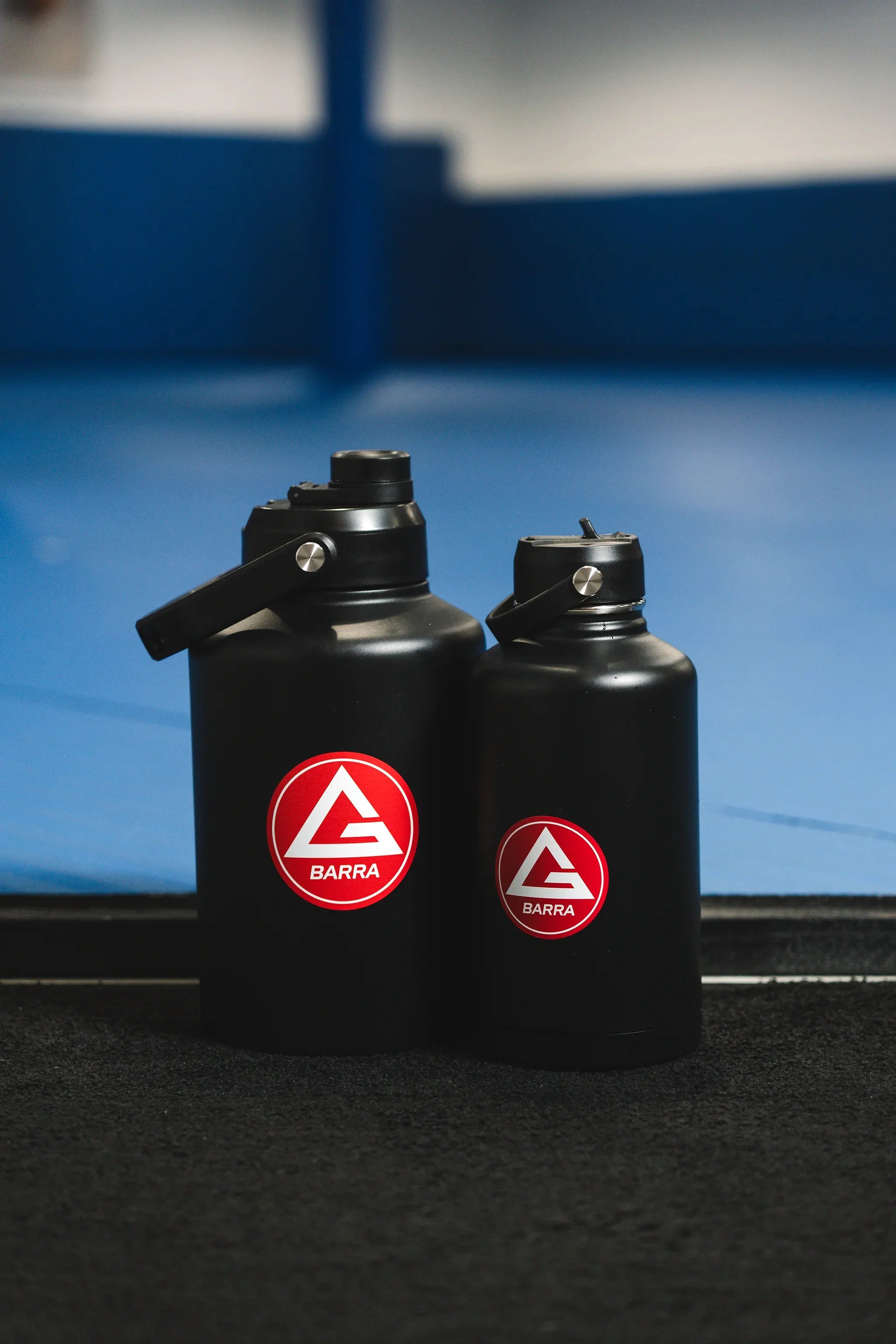 The black 128 oz and 64 oz Red Shield bottles with the Gracie Barra Red Shield logos, photographed side by side inside a GB training academy.