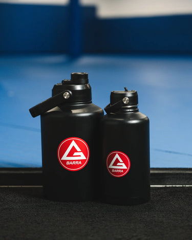 The black 128 oz and 64 oz Red Shield bottles with the Gracie Barra Red Shield logos, photographed side by side inside a GB training academy.
