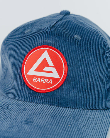 Close-up of the Red Shield Elite Cap's front panel showing the woven red Barra Shield patch on blue corduroy fabric, photographed on a white background.