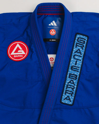 Close-up of blue Ripstop Lite Men’s BJJ kimono by adidas and Gracie Barra, featuring Red Shield chest patch, “Gracie Barra” embroidered lapel trim, and white adidas logo at the collar.