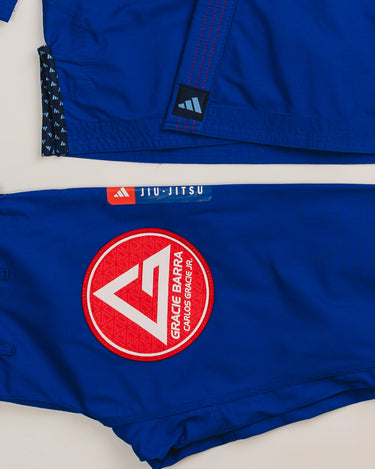 Close-up of blue Ripstop Lite Men’s BJJ kimono by adidas and Gracie Barra, showing blue adidas label on jacket hem, Red Shield patch on pants, and “Jiu-Jitsu” tag detail.