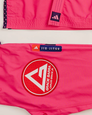 Close-up of pink Ripstop Lite Women’s BJJ kimono by adidas and Gracie Barra, showing the Red Shield patch on the pants, purple adidas label on the jacket hem, and “Jiu-Jitsu” tag detail.