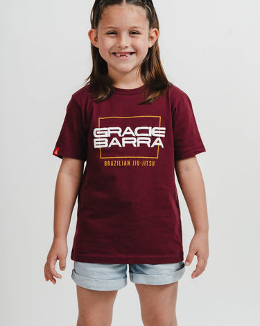 Young girl wearing maroon t-shirt with puff print "Gracie Barra Brazilian Jiu-Jitsu" logo, smiling against a white background.