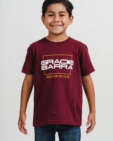 Young boy wearing maroon t-shirt with puff print "Gracie Barra Brazilian Jiu-Jitsu" logo and jeans, standing against a white background.
