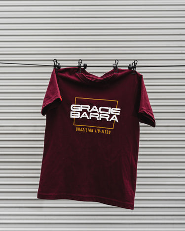 Maroon youth t-shirt with puff print "Gracie Barra Brazilian Jiu-Jitsu" logo, hanging on a clothesline against a metal background.