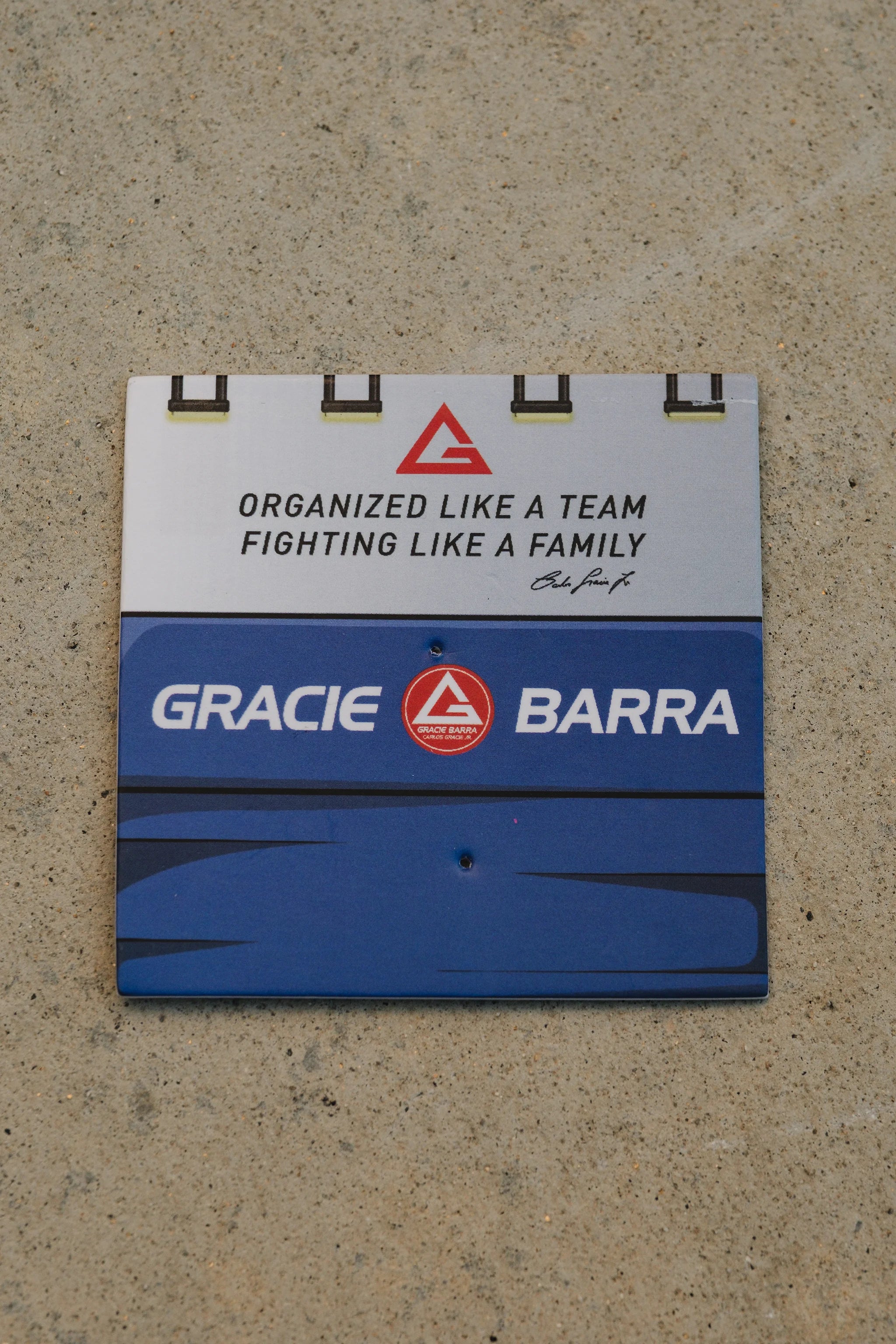 Gracie Barra branded backing card with “Organized Like a Team, Fighting Like a Family” text on a concrete surface.