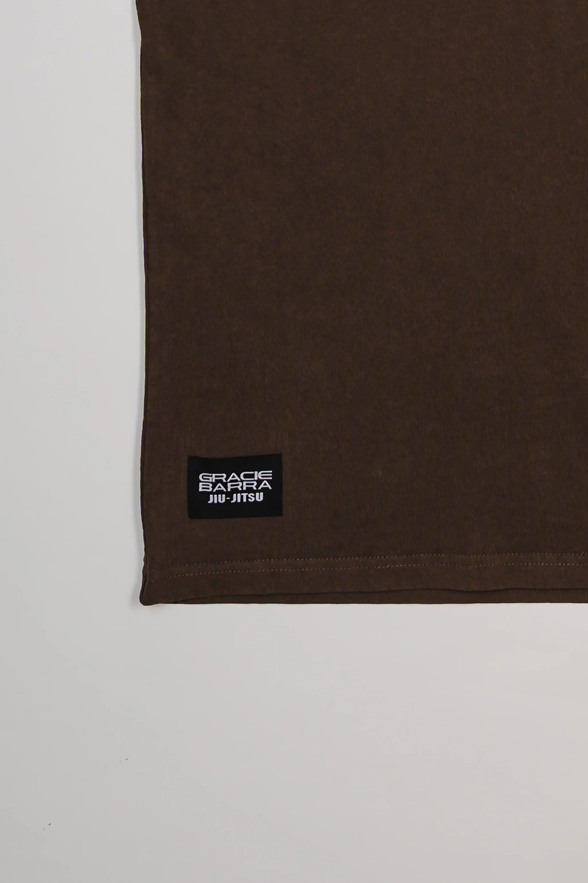 Close-up of bottom hem on Tactical Men’s Tee in dark brown with black woven Gracie Barra Jiu-Jitsu label, displayed on a light gray background.