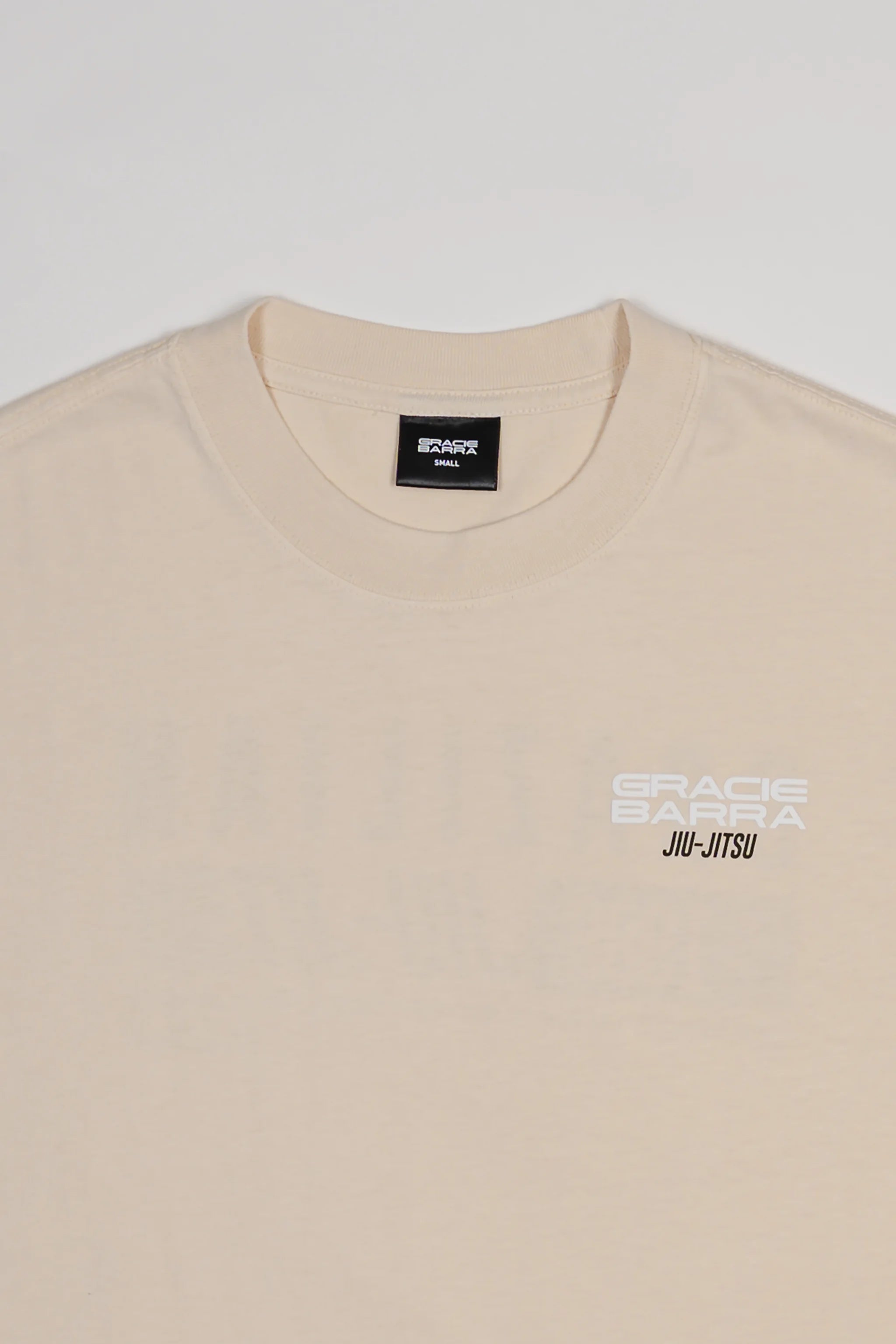 Close-up of chest area on Tactical Men’s Tee in natural beige with white Gracie Barra Jiu-Jitsu logo, displayed on a light gray background.
