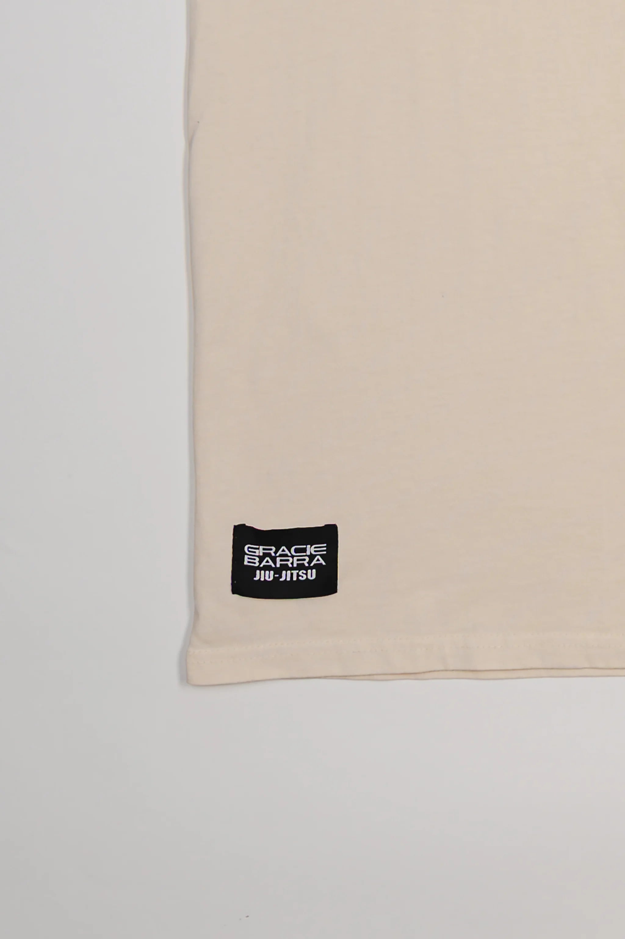 Close-up of bottom hem on Tactical Men’s Tee in natural beige featuring black woven Gracie Barra Jiu-Jitsu label, displayed on a light gray background.