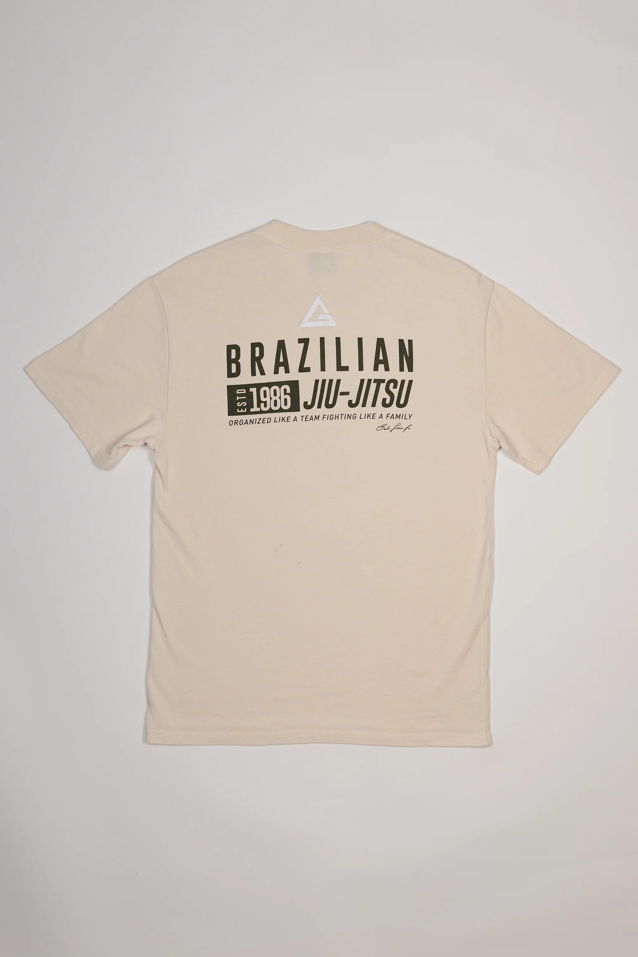 Back view of Tactical Men’s Tee in natural beige with Legacy G logo above bold “Brazilian Jiu-Jitsu” text and 1986 team heritage graphic, displayed on a light gray background.
