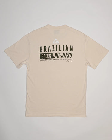 Back view of Tactical Men’s Tee in natural beige with Legacy G logo above bold “Brazilian Jiu-Jitsu” text and 1986 team heritage graphic, displayed on a light gray background.
