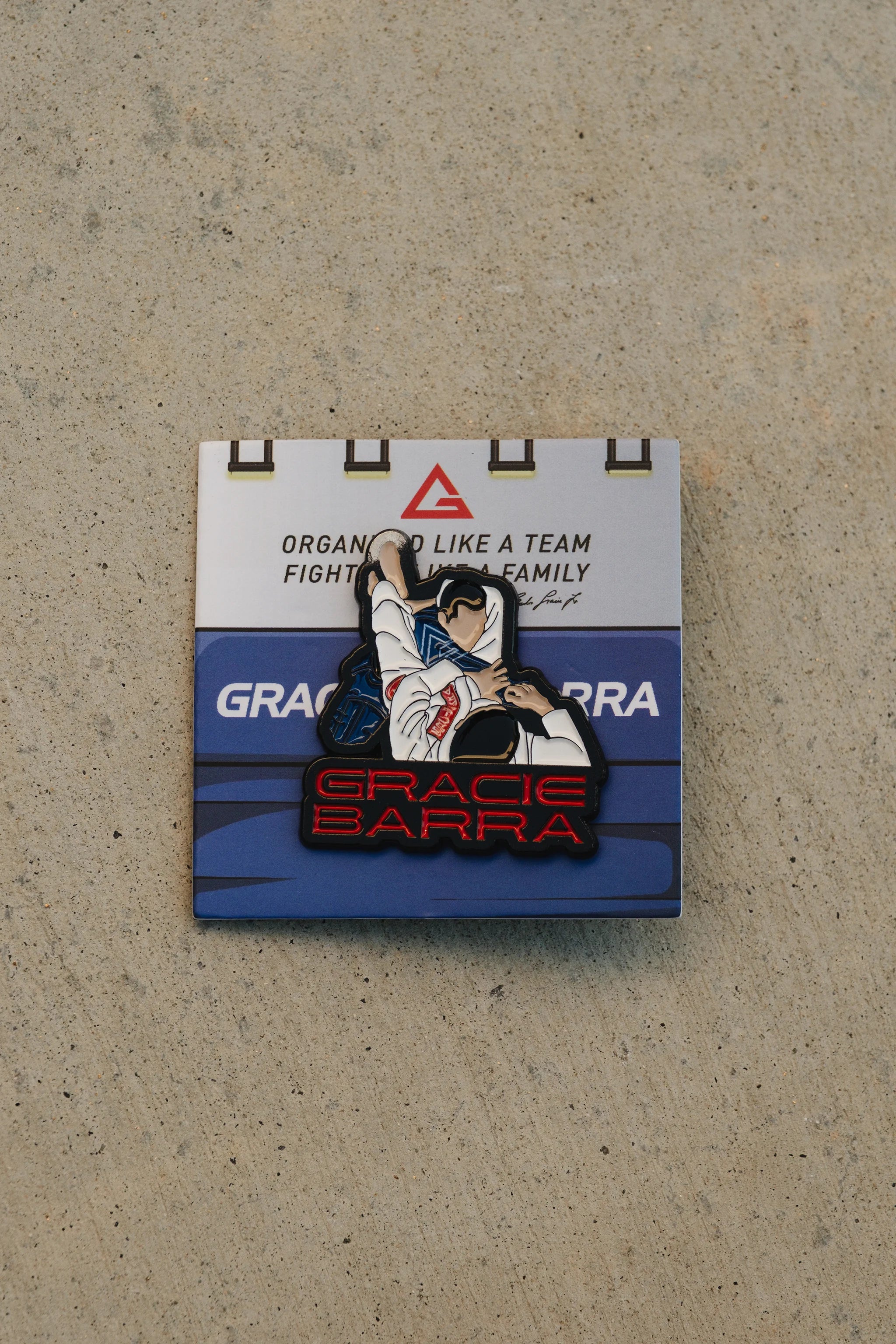 Gracie Barra triangle submission enamel pin mounted on a blue-and-white backing card with GB branding, photographed on a textured concrete surface.