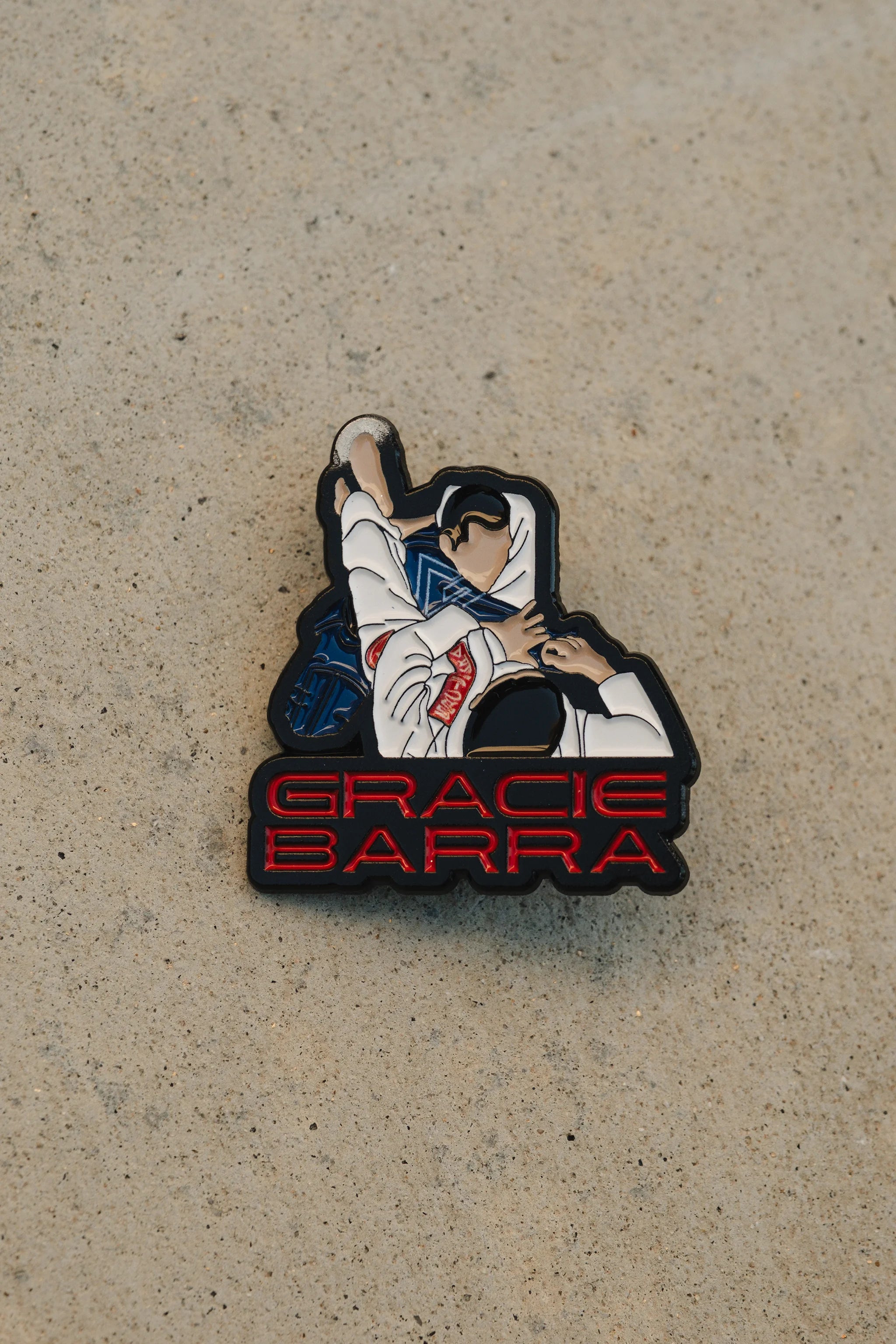 Close-up of the Triangle Pin showing the grappling artwork and red Gracie Barra lettering against a beige concrete background.