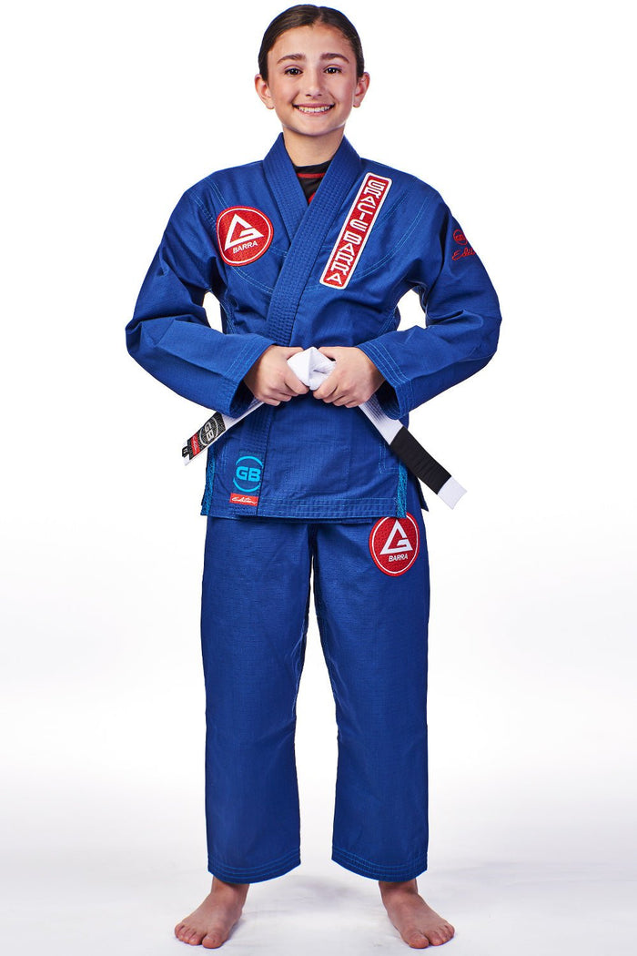 Child wearing the blue GBK Lite Ripstop kimono with a Red Shield chest patch, vertical Gracie Barra lapel patch, reinforced collar stitching, and a GB logo label at the bottom of the lapel. Pants feature a Red Shield patch on the thigh, and the white belt is tied at the waist