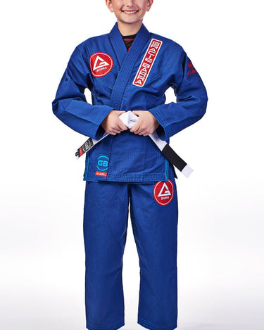 Child wearing the blue GBK Lite Ripstop kimono with a Red Shield chest patch, vertical Gracie Barra lapel patch, reinforced collar stitching, and a GB logo label at the bottom of the lapel. Pants feature a Red Shield patch on the thigh, and the white belt is tied at the waist