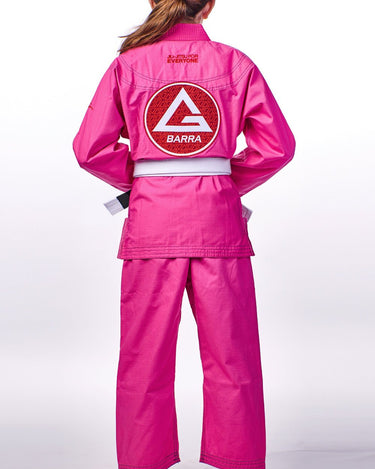 Back view of a young girl wearing the pink GBK Lite Ripstop kimono with a large Red Shield patch, reinforced shoulder panels, and a tied white belt—showing a comfortable, confident fit for training.